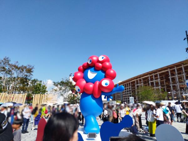 屋外広場に立つ2025年大阪・関西万博の公式マスコットミャクミャクの巨大な像。人混みの中にあります。