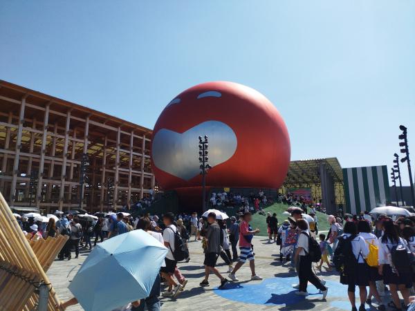 晴れた日、笑顔が書かれた巨大なオレンジのバルーンと、大勢の来場者が広場を歩く様子。