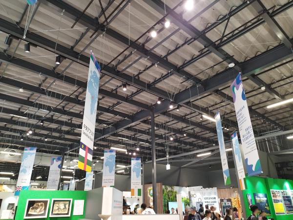 広い会場の天井から、世界地図と国旗がデザインされた複数の細長いバナーが吊り下げられています。