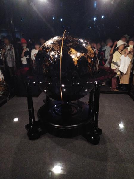 黒い台座に乗せられた、金色の模様が輝く黒い地球儀の展示品です。周囲に多くの人が立っています。