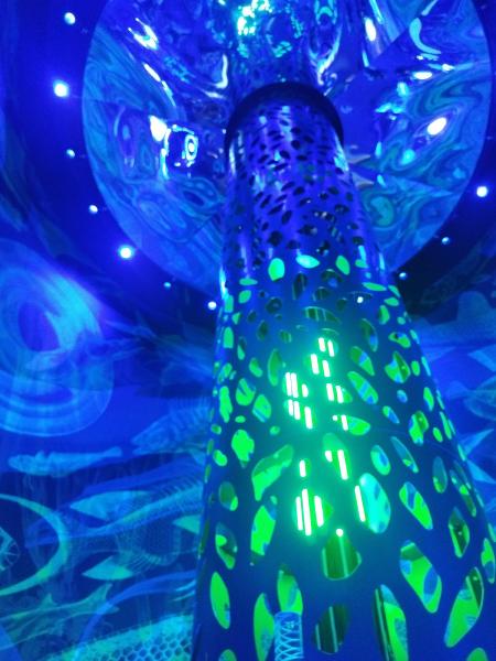 青と緑の光で照らされた、穴あきデザインの円柱の展示物。水中をイメージした青い空間です。