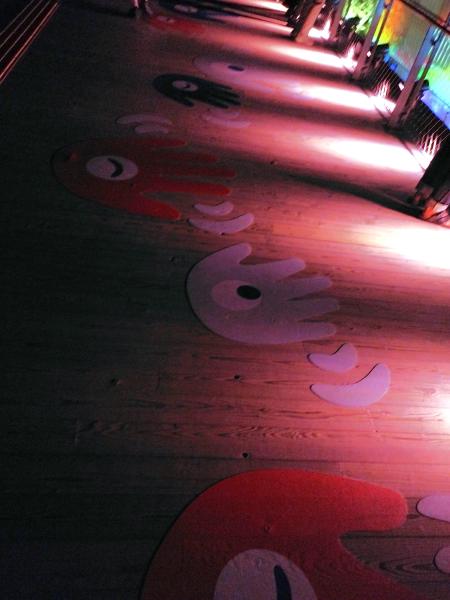 赤く照らされた木の床に、ミャクミャクをモチーフにした手と目の形の足跡風の模様が続く通路。