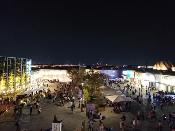 夜間の広い広場に、多くの人が集まり、周囲のパビリオンや店舗が明るく照らされています。