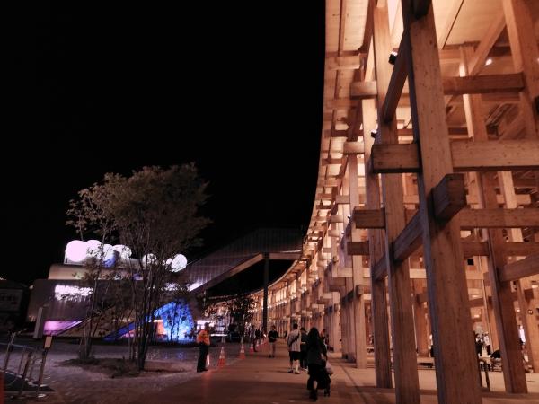 夜間、明るく照らされた巨大な木造リングと、遠くに見える小さなパビリオン。