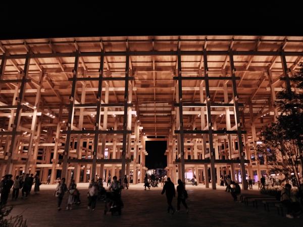 夜間の万博会場。明るく照らされた巨大な木組みの建物の下を人々が歩いています。