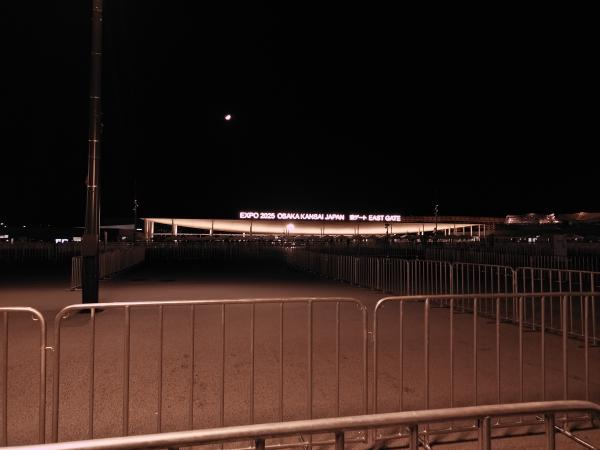 夜の大阪・関西万博の東ゲート。遠くに光る看板と、手前に並ぶ無人の規制柵。