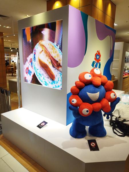 展示スペースに、食べ物の映像が流れるモニターと、ミャクミャクの大きなぬいぐるみ。