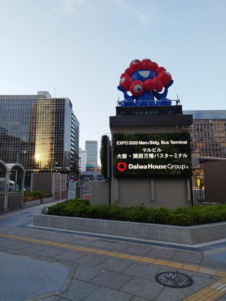 大阪駅近くの「EXPO 2025 Maru Bldg. Bus Terminal」を示す屋外看板です。看板の上には、万博公式キャラクターのミャクミャクの巨大なオブジェが設置されており、街中で万博をPRしています。