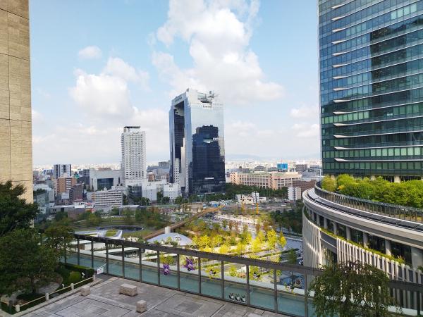 大阪ステーションシティの風の広場からの眺めです。高層ビルが立ち並ぶ大阪の都市景観を上層階から捉えています。中央には、独特な形をした梅田スカイビルが見え、手前には緑豊かな公園や建物の屋上庭園が広がっています。