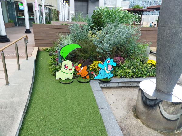 建物の屋外広場にある花壇の前で、チコリータ、ヒノアラシ、ワニノコの等身大パネルが並んでいます。背景の植え込みの緑とキャラクターが調和しています。