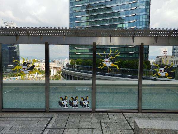 建物の屋外広場にあるガラスの仕切りに、黄色と黒の電気タイプのポケモン、エモンガのパネルが飾られています。遠景には都会の街並みが見えます。
