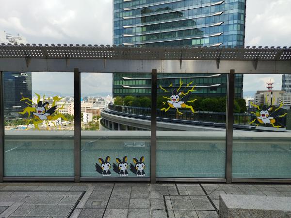 建物の屋外広場にあるガラスの仕切りに、黄色と黒の電気タイプのポケモン、エモンガのパネルが飾られています。遠景には都会の街並みが見えます。