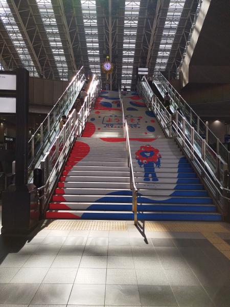 駅構内の大きな階段に、2025年大阪・関西万博のミャクミャクのイラストと赤と青の模様が描かれています。上部にはシンボルの金色の大きな時計が見えます。