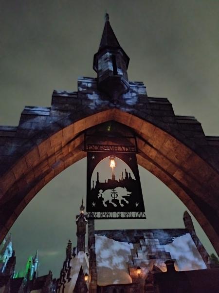 石造りの門の上部。中央に「HOGSMEADE」の旗。夜空の下、ライトアップされている。