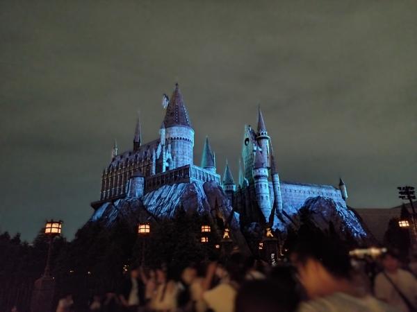 夜間に青い光で照らされたホグワーツ城。手前に多くの人々の後頭部が見える。