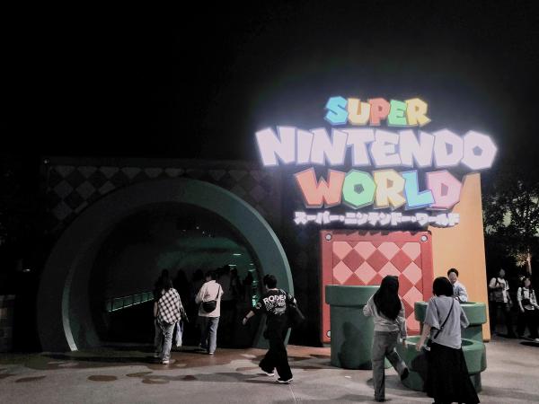「SUPER NINTENDO WORLD」の看板が光る入り口。緑の土管型のトンネルを人々が歩いている。